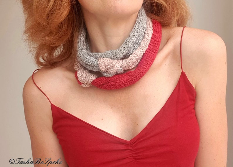 May include: A knitted infinity scarf in shades of gray, pink, and red. The scarf is twisted and draped around the neck. The model is wearing a red spaghetti strap top.