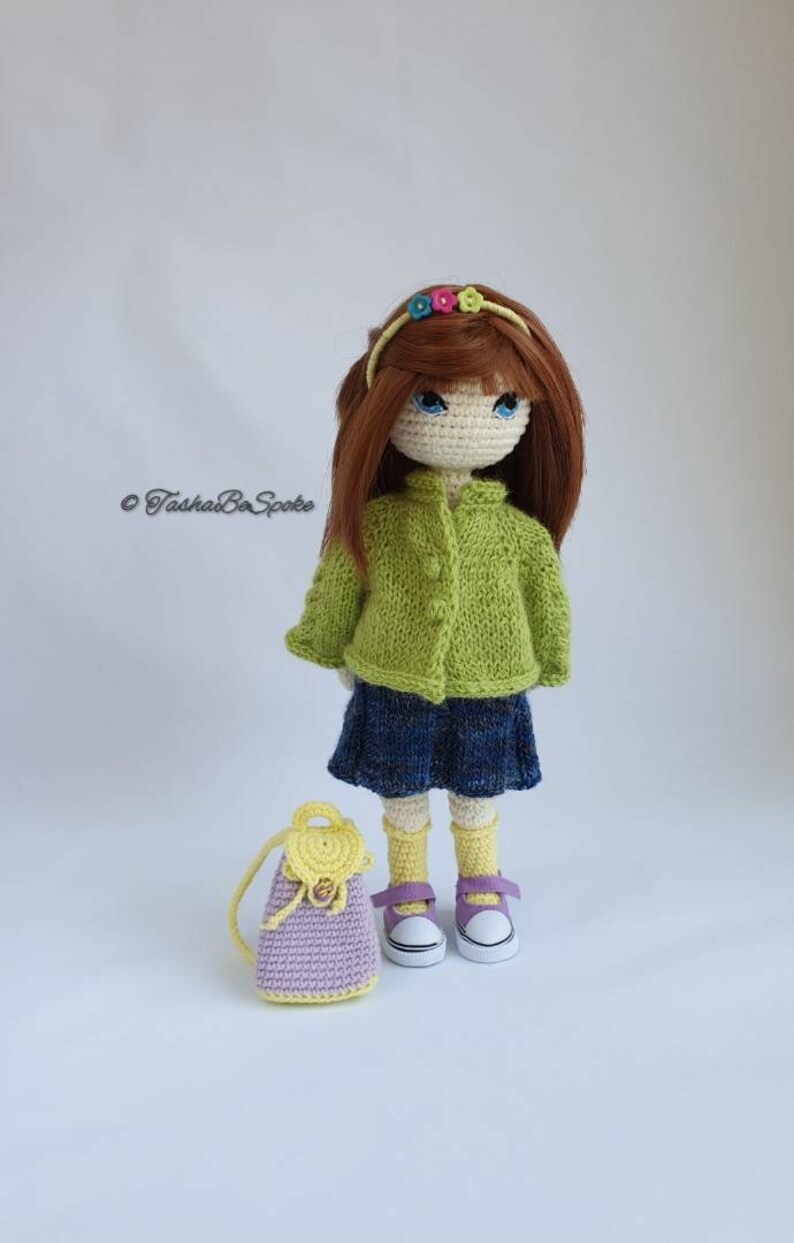 Pu&ograve; includere: Una bambola all'uncinetto che indossa un cardigan verde, una gonna blu e calzini gialli. La bambola ha i capelli castani, gli occhi azzurri e una fascia per capelli con fiori colorati. La bambola tiene uno zaino all'uncinetto viola e giallo.