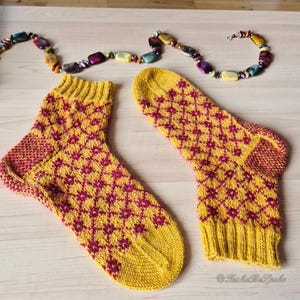 May include: A pair of yellow knitted socks with a repeating burgundy diamond pattern. The socks have ribbed cuffs and heels. A colorful beaded necklace is above the socks on a light wooden surface.