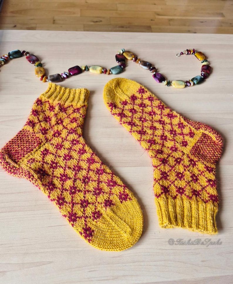 May include: A pair of yellow knitted socks with a repeating burgundy diamond pattern. The socks have ribbed cuffs and heels. A colorful beaded necklace is above the socks on a light wooden surface.
