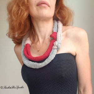 May include: A knitted necklace in shades of gray and red. The necklace is knotted in the front and appears to be made of a soft, chunky yarn. The design is a unique accessory.