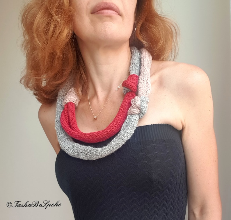 May include: A knitted necklace in shades of gray and red. The necklace is knotted in the front and appears to be made of a soft, chunky yarn. The design is a unique accessory.