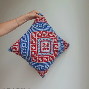 Op de afbeelding: Een vierkant gehaakt kussen met een geometrisch ontwerp in rood, blauw en lichtblauw. Het kussen heeft een centraal vierkant met een stervormig patroon, omgeven door herhaalde vierkante en rechthoekige motieven. Het kussen wordt door een hand vastgehouden.