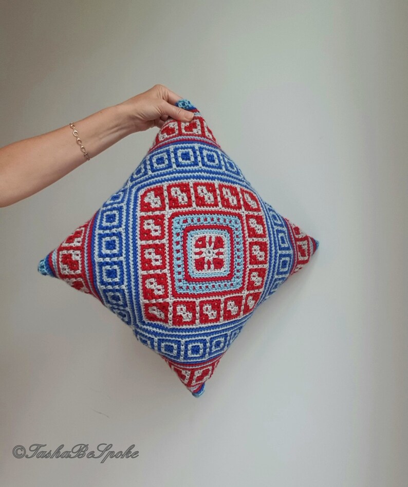 Op de afbeelding: Een vierkant gehaakt kussen met een geometrisch ontwerp in rood, blauw en lichtblauw. Het kussen heeft een centraal vierkant met een stervormig patroon, omgeven door herhaalde vierkante en rechthoekige motieven. Het kussen wordt door een hand vastgehouden.