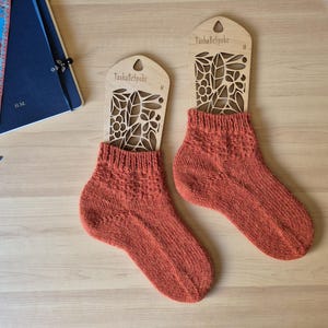 May include: Two pairs of rust-colored knitted socks displayed on wooden sock forms. The forms are laser-cut with a floral design and the text "TashaBeSpoke". The socks have a textured pattern and are presented on a light wooden surface.