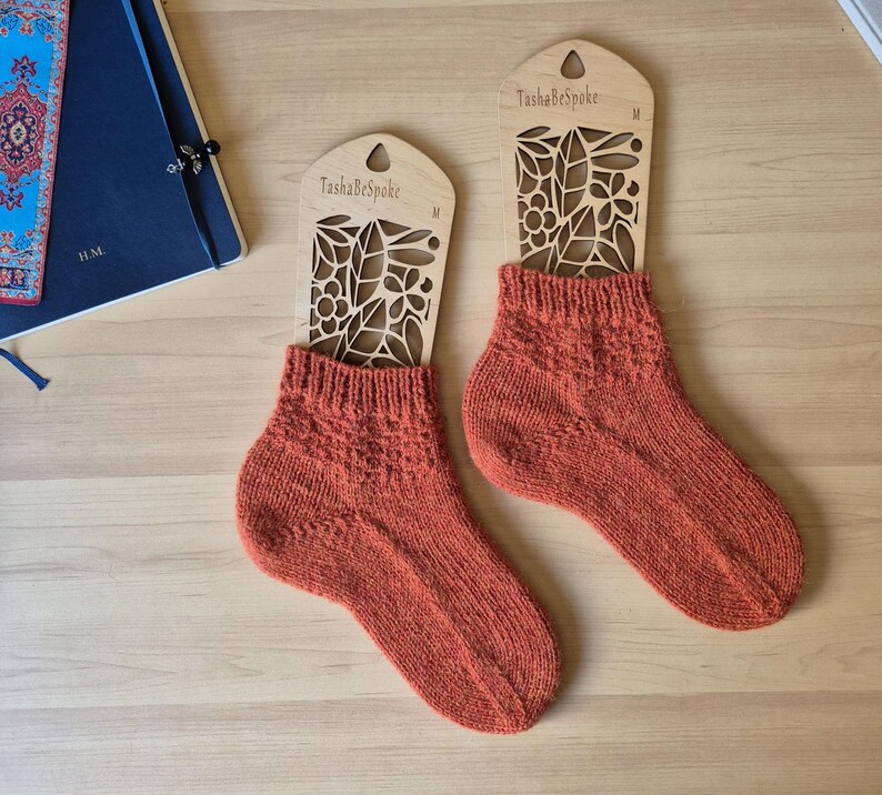 May include: Two pairs of rust-colored knitted socks displayed on wooden sock forms. The forms are laser-cut with a floral design and the text "TashaBeSpoke". The socks have a textured pattern and are presented on a light wooden surface.