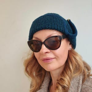 K&ouml;nnte beinhalten: Eine Frau tr&auml;gt eine tealblaue Strickm&uuml;tze mit umgeklapptem Rand und eine dunkle Sonnenbrille. Sie tr&auml;gt einen braunen Mantel mit einer dekorativen Brosche.