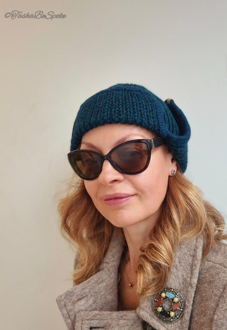 K&ouml;nnte beinhalten: Eine Frau tr&auml;gt eine tealblaue Strickm&uuml;tze mit umgeklapptem Rand und eine dunkle Sonnenbrille. Sie tr&auml;gt einen braunen Mantel mit einer dekorativen Brosche.