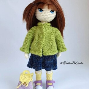 Pu&ograve; includere: Una bambola all'uncinetto con i capelli castani, che indossa un cardigan verde, una gonna blu, calzini gialli e scarpe viola. La bambola tiene uno zaino all'uncinetto viola e giallo.