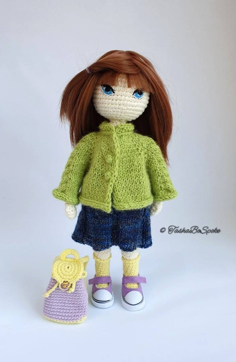 Pu&ograve; includere: Una bambola all'uncinetto con i capelli castani, che indossa un cardigan verde, una gonna blu, calzini gialli e scarpe viola. La bambola tiene uno zaino all'uncinetto viola e giallo.