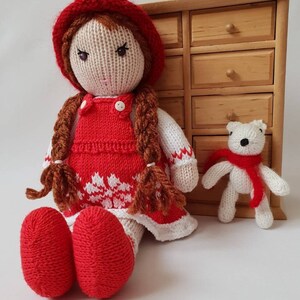 May include: A knitted doll with red hair, a red dress, and a red hat. The doll is sitting next to a small white knitted bear wearing a red scarf. The doll and bear are sitting in front of a wooden dresser with drawers.