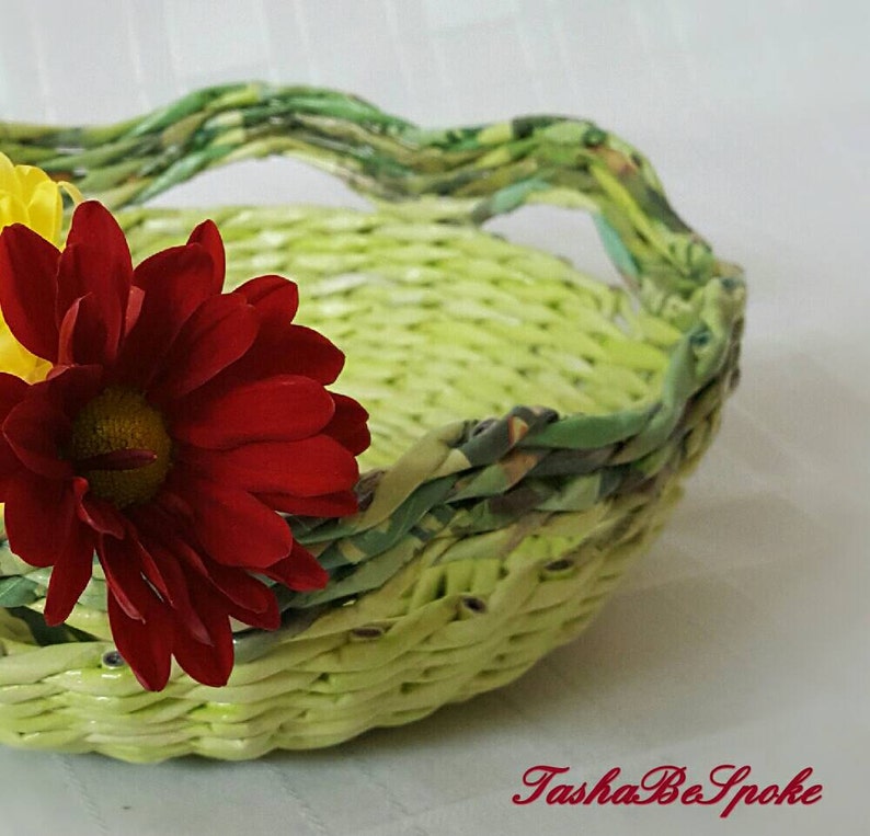 Puede incluir: Una cesta tejida a mano, en tonos verdes y amarillos, con asa. Una vibrante flor roja y una flor amarilla se colocan en el lateral de la cesta. La cesta probablemente se utiliza con fines decorativos.