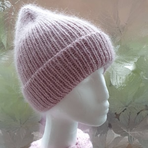 K&ouml;nnte beinhalten: Eine rosafarbene Strickm&uuml;tze mit flauschigem Muster. Die M&uuml;tze sitzt auf einem wei&szlig;en Mannequin-Kopf. Der Hintergrund ist ein verschwommenes Bild von Bl&auml;ttern.