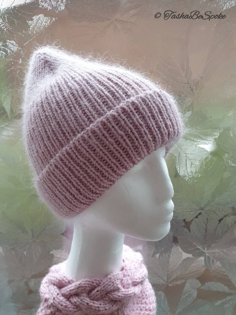K&ouml;nnte beinhalten: Eine rosafarbene Strickm&uuml;tze mit flauschigem Muster. Die M&uuml;tze sitzt auf einem wei&szlig;en Mannequin-Kopf. Der Hintergrund ist ein verschwommenes Bild von Bl&auml;ttern.
