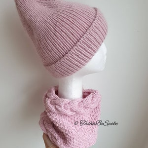 K&ouml;nnte beinhalten: Eine rosa gestrickte M&uuml;tze und ein passender Infinity-Schal mit Zopfmuster. Der Schal wird in einer Hand mit einem goldenen Armband gehalten.