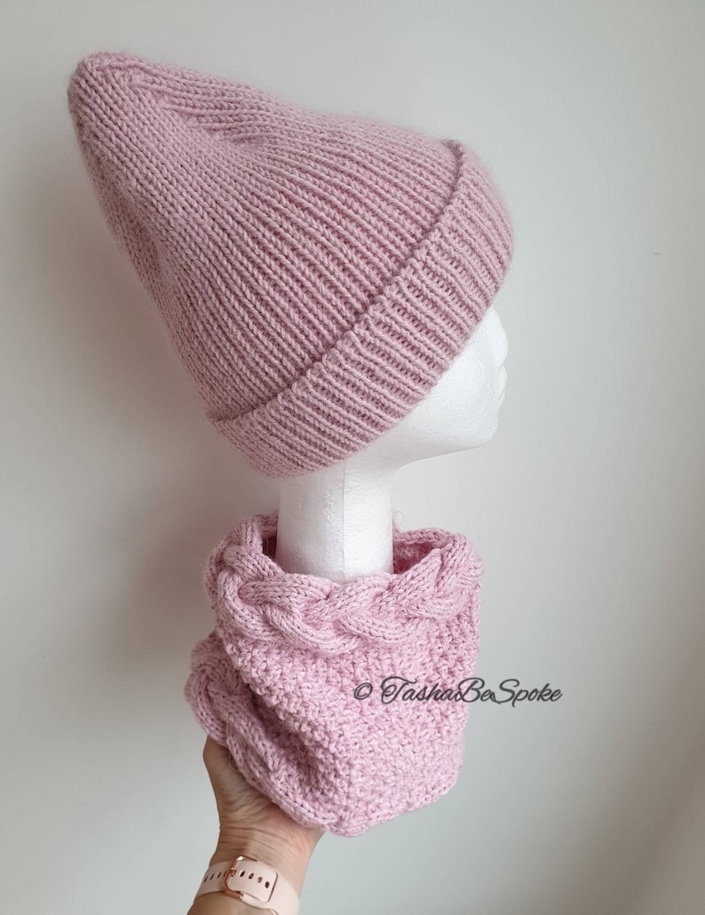 K&ouml;nnte beinhalten: Eine rosa gestrickte M&uuml;tze und ein passender Infinity-Schal mit Zopfmuster. Der Schal wird in einer Hand mit einem goldenen Armband gehalten.