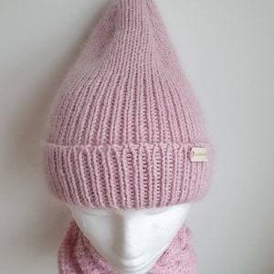 K&ouml;nnte beinhalten: Eine rosafarbene Strickm&uuml;tze und ein passender Schal. Die M&uuml;tze hat eine spitze Krone und der Schal ist ein Schlauchschal. Die M&uuml;tze hat ein kleines Etikett mit der Aufschrift "TashaBeSpoke".