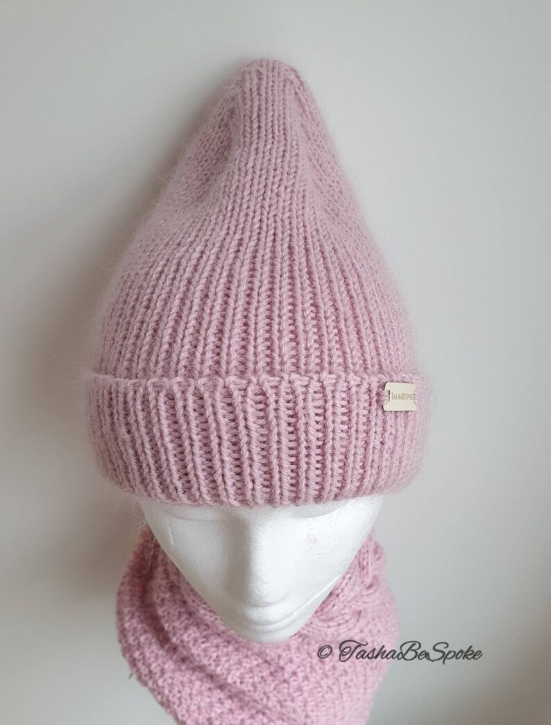 K&ouml;nnte beinhalten: Eine rosafarbene Strickm&uuml;tze und ein passender Schal. Die M&uuml;tze hat eine spitze Krone und der Schal ist ein Schlauchschal. Die M&uuml;tze hat ein kleines Etikett mit der Aufschrift "TashaBeSpoke".
