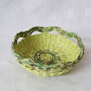 Puede incluir: Una cesta redonda, tejida a mano, con una combinaci&oacute;n de colores verde claro y verde oliva. La cesta est&aacute; hecha de tiras de papel tejidas apretadamente, creando una superficie texturizada. El borde tiene un dise&ntilde;o ondulado. Esta cesta decorativa es adecuada para la decoraci&oacute;n del hogar.