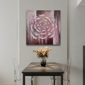 May include: Textured canvas art depicting a white and pink rose. The artwork is set against a backdrop of vertical stripes in shades of brown and pink. The piece is displayed above a table with a vase of dried flowers.