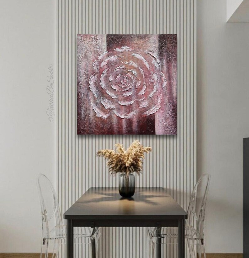 May include: Textured canvas art depicting a white and pink rose. The artwork is set against a backdrop of vertical stripes in shades of brown and pink. The piece is displayed above a table with a vase of dried flowers.