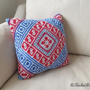 Op de afbeelding: Vierkant decoratief kussen met een geometrisch patroon in rood, wit en blauw. Het kussen is gehaakt met een gedetailleerd ontwerp, met ruit- en vierkantvormen. Het kussen ligt op een beige bank.