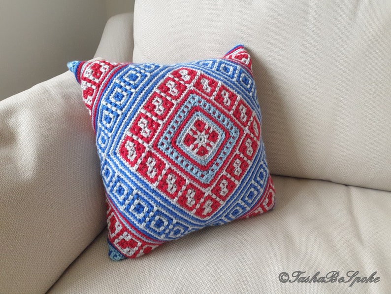 Op de afbeelding: Vierkant decoratief kussen met een geometrisch patroon in rood, wit en blauw. Het kussen is gehaakt met een gedetailleerd ontwerp, met ruit- en vierkantvormen. Het kussen ligt op een beige bank.