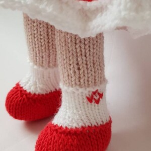 May include: Close-up of a knitted doll's feet wearing red and white knitted shoes with a white knitted cuff. The shoes have a red embroidered design on the cuff.