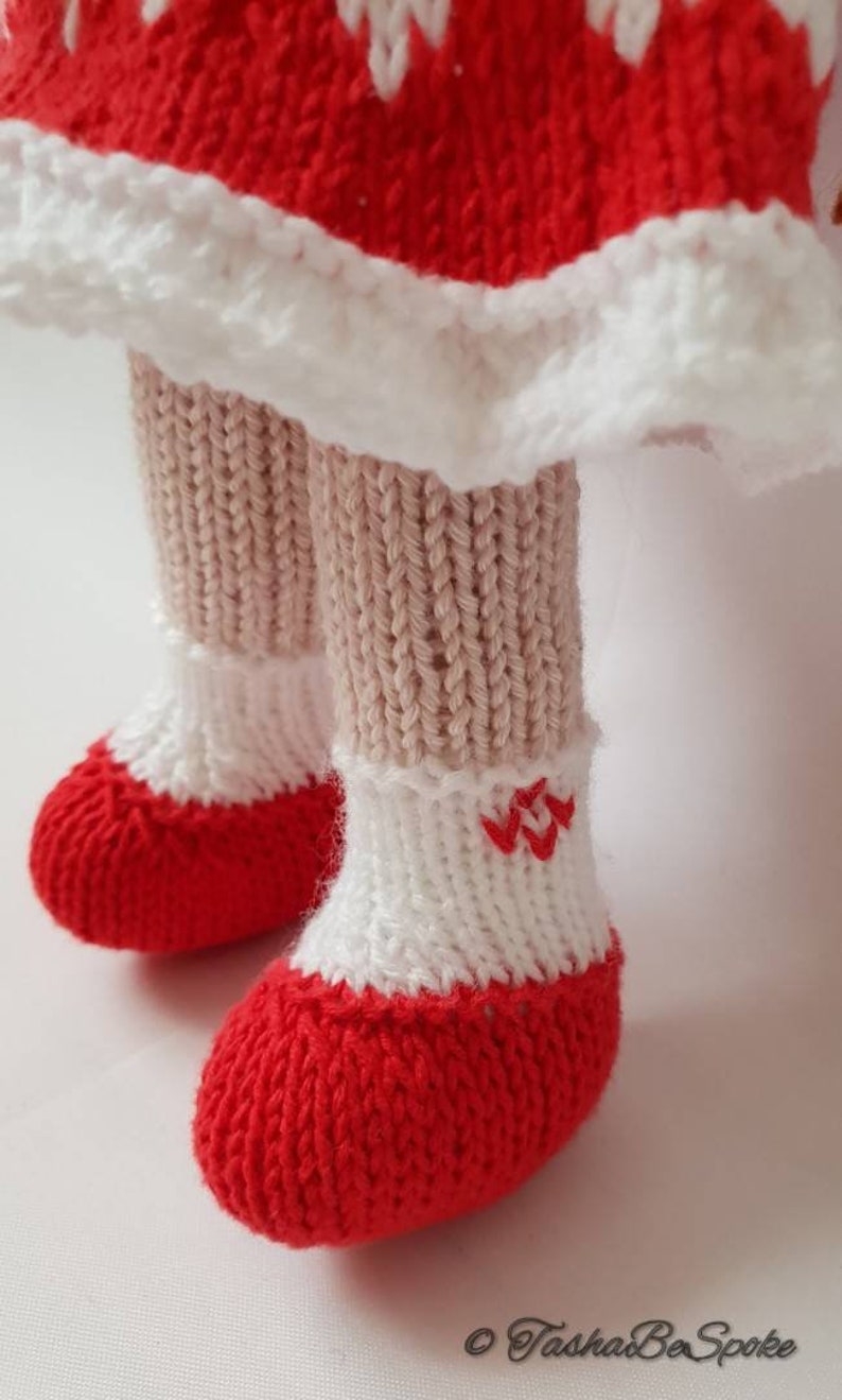 May include: Close-up of a knitted doll's feet wearing red and white knitted shoes with a white knitted cuff. The shoes have a red embroidered design on the cuff.