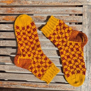 May include: A pair of knitted socks in a golden yellow color with a repeating pattern of dark red diamond shapes. The heel and toe sections are a darker shade of orange-red. The socks are displayed on a wooden surface.
