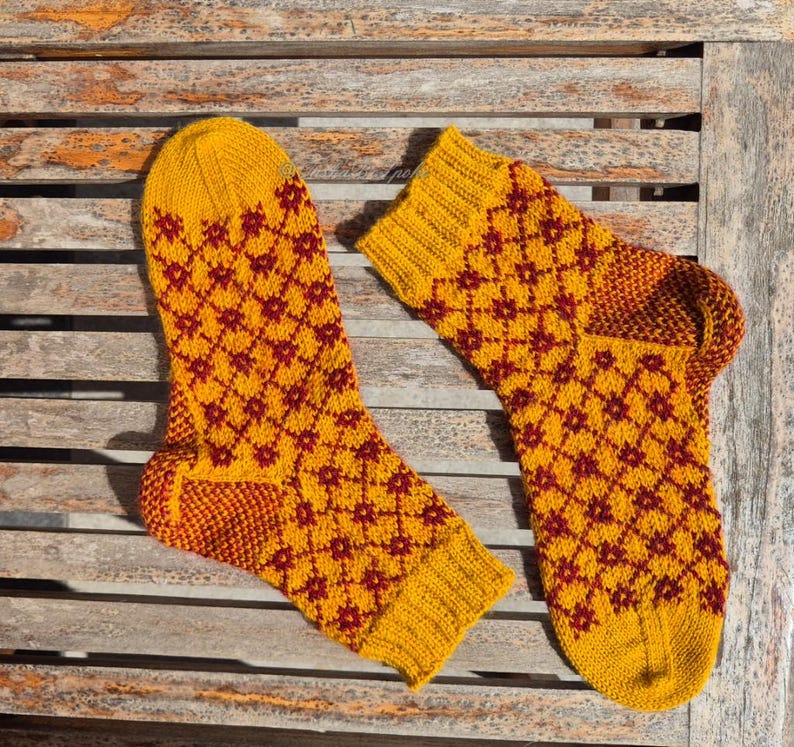 May include: A pair of knitted socks in a golden yellow color with a repeating pattern of dark red diamond shapes. The heel and toe sections are a darker shade of orange-red. The socks are displayed on a wooden surface.