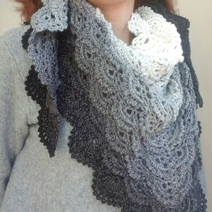 May include: A black, grey, and white crocheted shawl with a scalloped edge. The shawl is made with a gradient yarn that transitions from black to grey to white. The shawl is worn by a person with red hair.