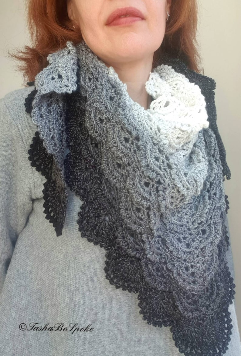 May include: A black, grey, and white crocheted shawl with a scalloped edge. The shawl is made with a gradient yarn that transitions from black to grey to white. The shawl is worn by a person with red hair.