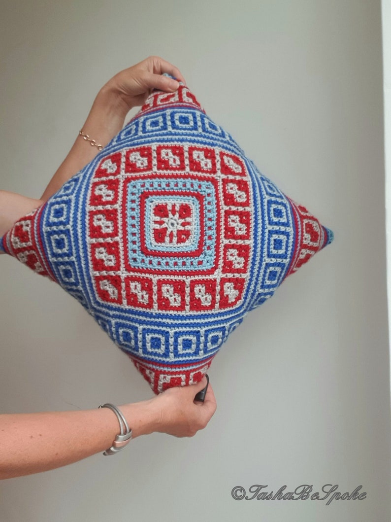 Op de afbeelding: Een vierkant, gehaakt kussen met een geometrisch ontwerp in rood, blauw en wit. Het kussen heeft een centraal vierkant met een bloemachtig patroon, omgeven door herhalende vierkante en rechthoekige vormen. Het kussen wordt door twee handen omhoog gehouden.