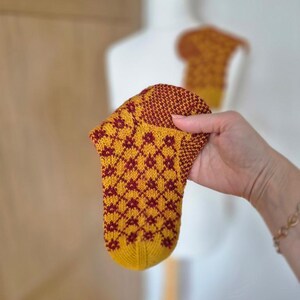 May include: A hand-knitted sock with a yellow base and a repeating maroon floral pattern. The sock is held up, showcasing its intricate design and craftsmanship. A second sock is visible in the background.