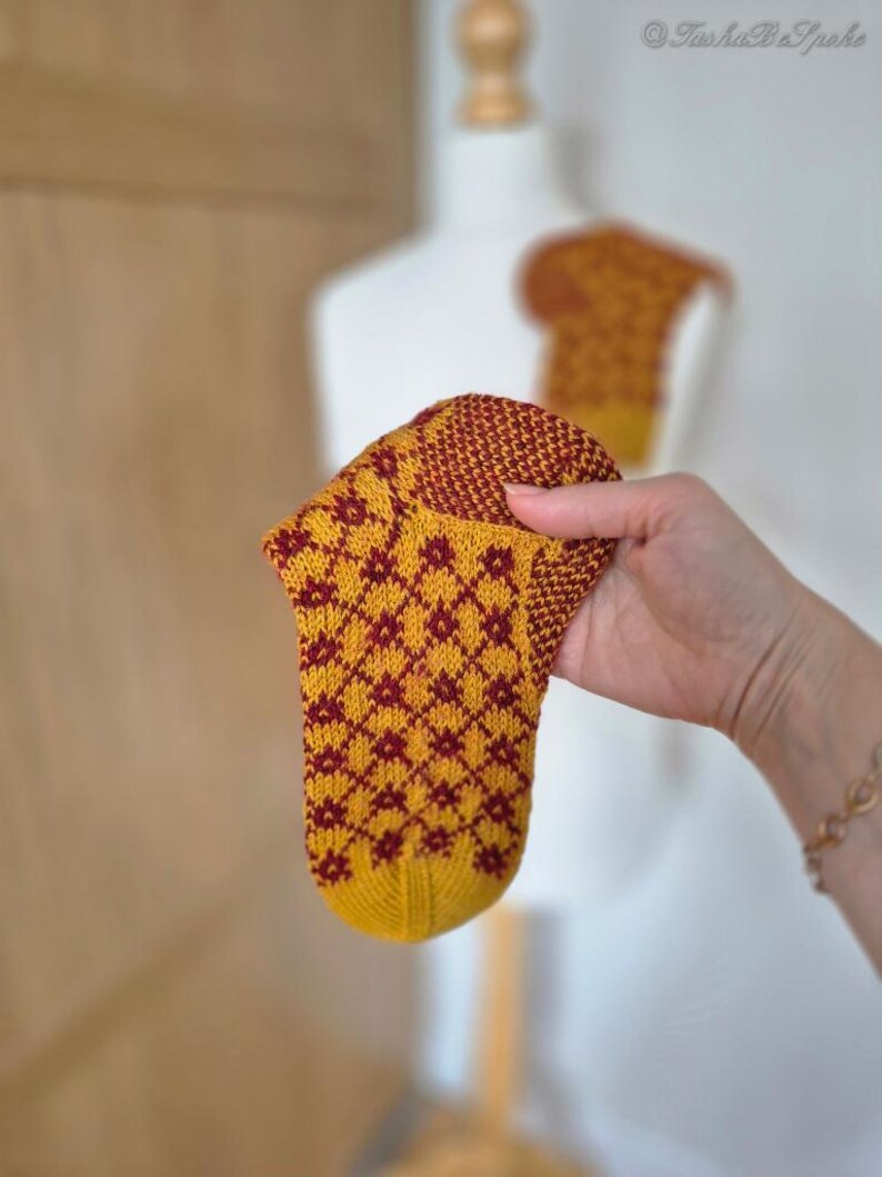 May include: A hand-knitted sock with a yellow base and a repeating maroon floral pattern. The sock is held up, showcasing its intricate design and craftsmanship. A second sock is visible in the background.