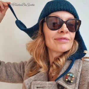K&ouml;nnte beinhalten: Eine tealblaue Strickm&uuml;tze mit einem langen Ohrenklappen. Die M&uuml;tze wird von einer Person mit braunen Haaren und Sonnenbrille getragen. Die Person tr&auml;gt einen braunen Mantel mit einer silbernen Brosche.