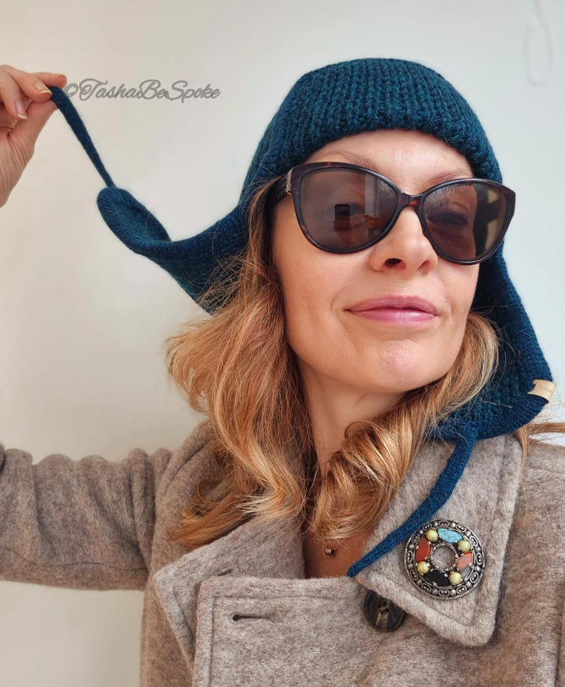 K&ouml;nnte beinhalten: Eine tealblaue Strickm&uuml;tze mit einem langen Ohrenklappen. Die M&uuml;tze wird von einer Person mit braunen Haaren und Sonnenbrille getragen. Die Person tr&auml;gt einen braunen Mantel mit einer silbernen Brosche.