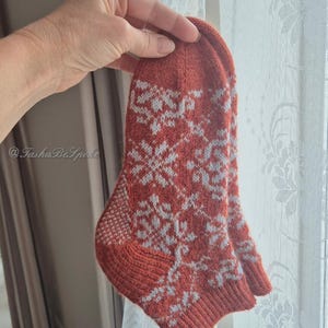 May include: A pair of hand-knitted socks in a burnt orange color with a white snowflake pattern. The socks are held up in front of a window. The cuff, heel, and toe are a solid orange color.