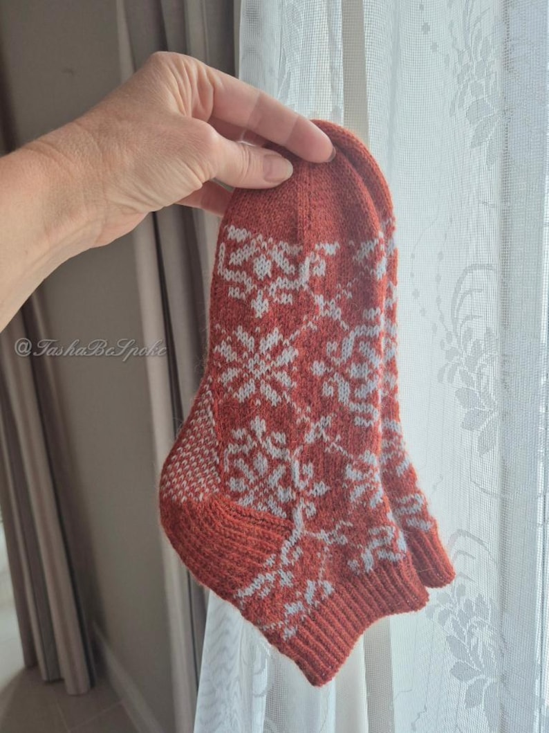 May include: A pair of hand-knitted socks in a burnt orange color with a white snowflake pattern. The socks are held up in front of a window. The cuff, heel, and toe are a solid orange color.