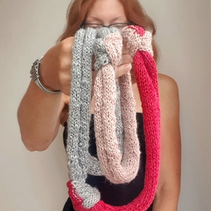 May include: A hand-knitted infinity scarf in three colors: gray, pink, and red. The scarf is held up in front of a person, showcasing the texture and color variations. The scarf is a loop design, perfect for adding warmth and style.