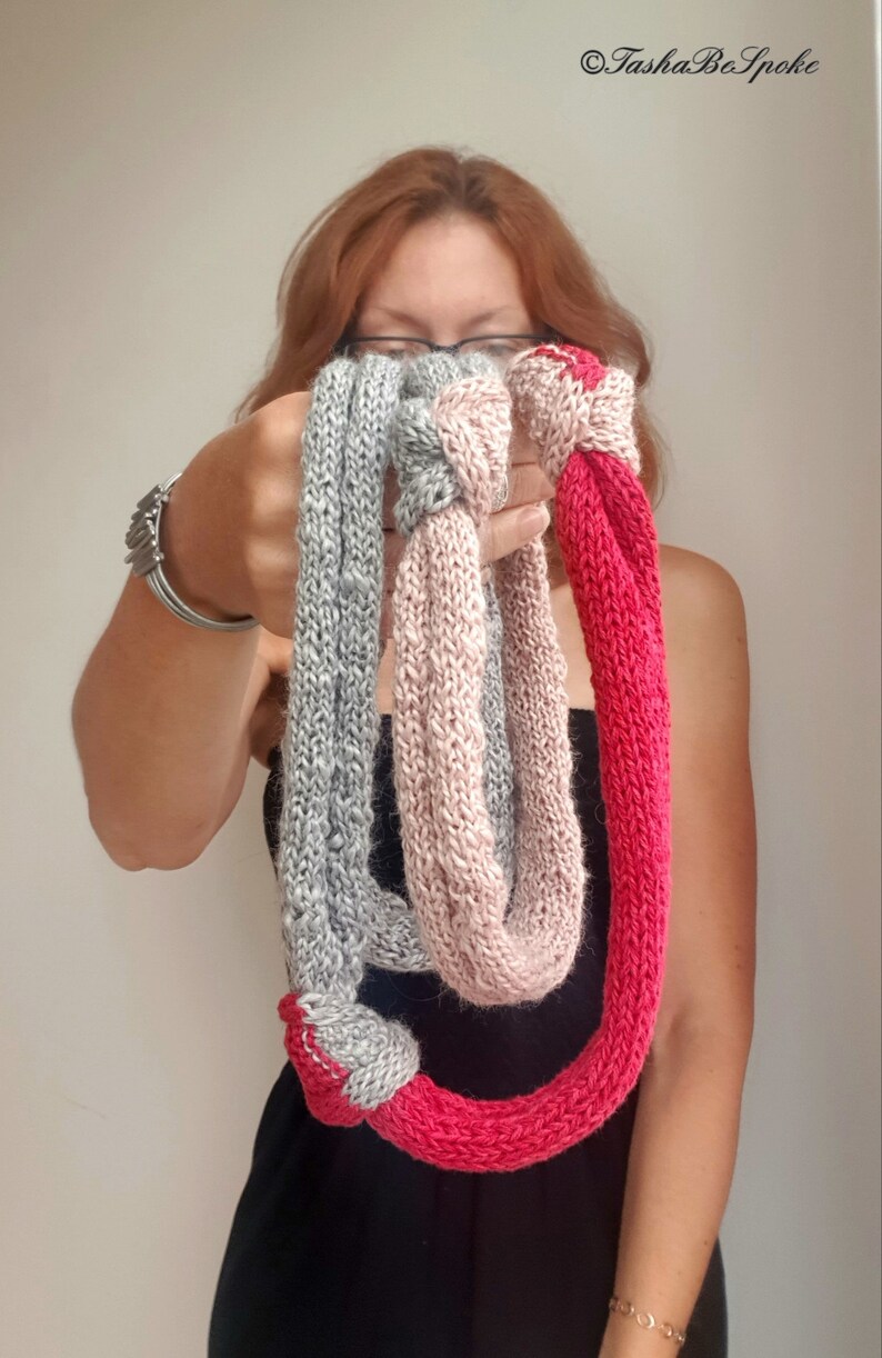 May include: A hand-knitted infinity scarf in three colors: gray, pink, and red. The scarf is held up in front of a person, showcasing the texture and color variations. The scarf is a loop design, perfect for adding warmth and style.