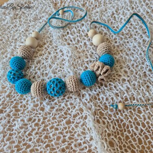 Op de afbeelding: Een handgemaakte ketting met gehaakte en houten kralen. De ketting heeft afwisselend lichtblauwe en beige gehaakte kralen, sommige met een open patroon. Het koord is blauw en wit, met houten kralen aan de uiteinden. De ketting is ongeveer 40 cm lang.