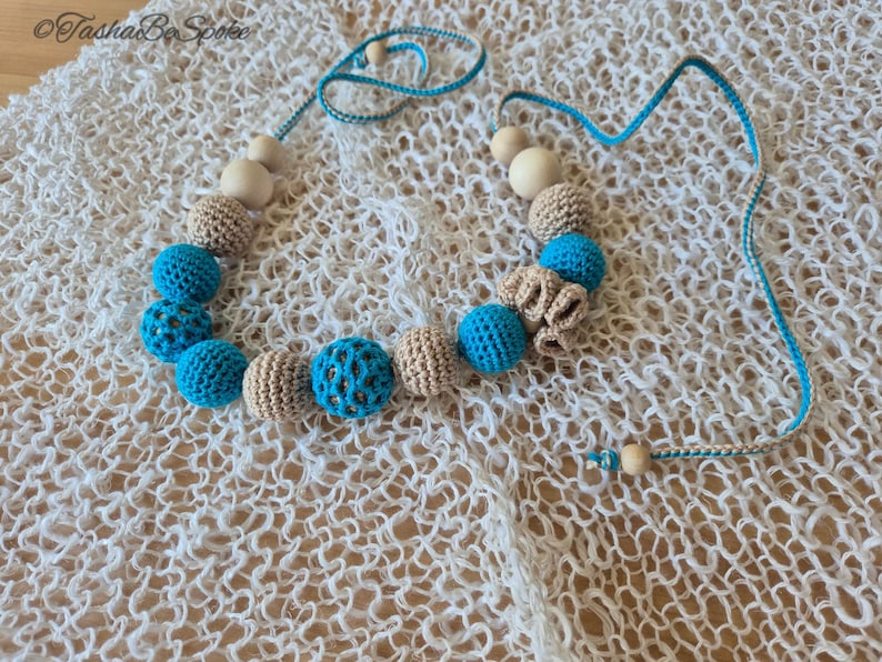 Op de afbeelding: Een handgemaakte ketting met gehaakte en houten kralen. De ketting heeft afwisselend lichtblauwe en beige gehaakte kralen, sommige met een open patroon. Het koord is blauw en wit, met houten kralen aan de uiteinden. De ketting is ongeveer 40 cm lang.