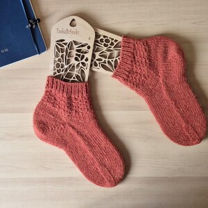 May include: A pair of coral-colored knitted socks displayed on a wooden sock form. The socks have a textured pattern on the cuff and top of the foot. The wooden form has a floral cut-out design and the text "TashaBespoke".