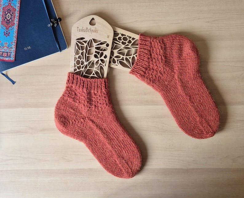 May include: A pair of coral-colored knitted socks displayed on a wooden sock form. The socks have a textured pattern on the cuff and top of the foot. The wooden form has a floral cut-out design and the text "TashaBespoke".