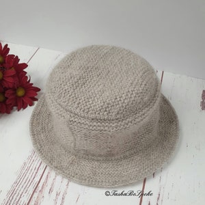 May include: A light beige knitted bucket hat with a rounded crown and a short brim. The hat is made of a soft, textured yarn. The hat is sitting on a light wood surface. Red flowers are in the background. The text "TashaBeSpoke" is on the bottom right.