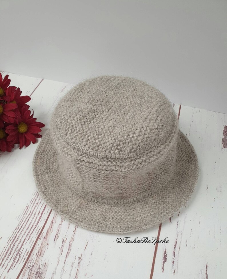May include: A light beige knitted bucket hat with a rounded crown and a short brim. The hat is made of a soft, textured yarn. The hat is sitting on a light wood surface. Red flowers are in the background. The text "TashaBeSpoke" is on the bottom right.