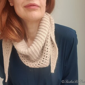 Puede incluir: Una bufanda de punto beige con un delicado patr&oacute;n de encaje. La bufanda est&aacute; drapeada alrededor del cuello de una persona que lleva una camisa azul oscuro.