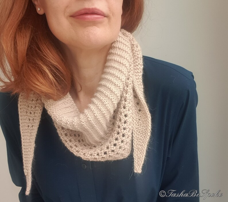Puede incluir: Una bufanda de punto beige con un delicado patr&oacute;n de encaje. La bufanda est&aacute; drapeada alrededor del cuello de una persona que lleva una camisa azul oscuro.